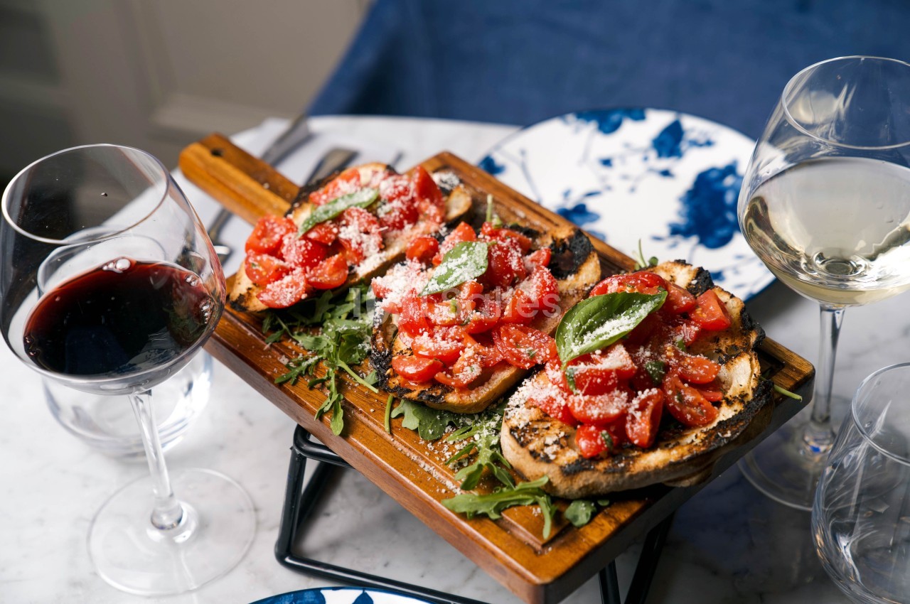 Tranches de pain grillé garnies de tomates, basilic et fromage, servies sur un plateau en bois avec des verres de vin.