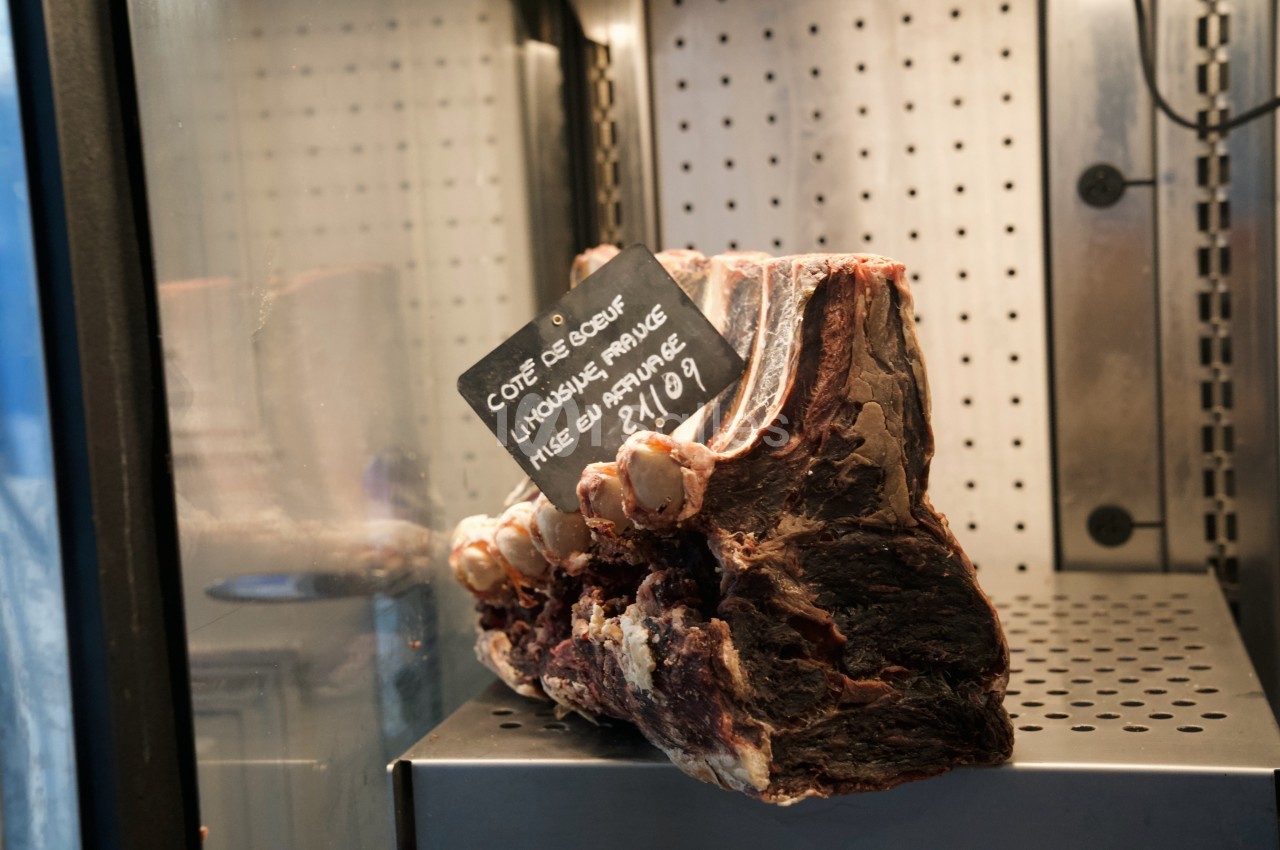 Pièce de côte de bœuf maturée exposée dans une vitrine réfrigérée avec une étiquette indiquant son origine et son prix.