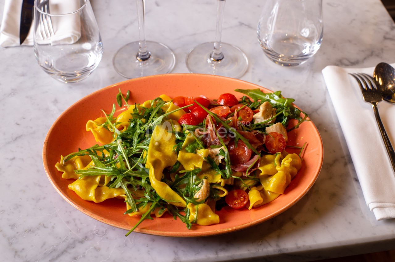 Assiette de pâtes fraîches garnies de roquette, tomates cerises et jambon cru, servie sur une table en marbre.