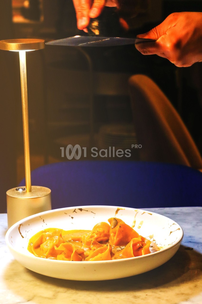 Assiette de raviolis servis sur une table en marbre, éclairée par une lampe, avec des mains ajustant un élément au-dessus.