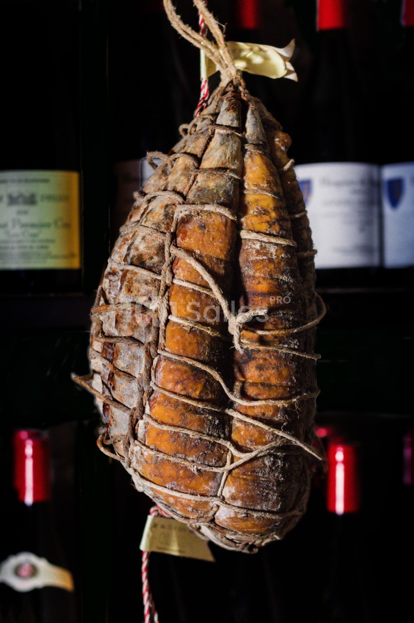 Saucisson sec artisanal suspendu dans un filet devant des bouteilles de vin en arrière-plan flou.
