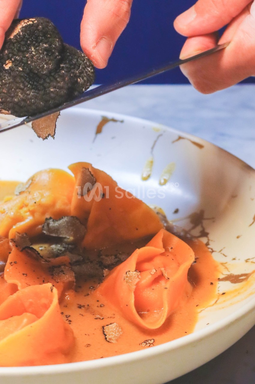 Des mains râpent une truffe noire sur des pâtes fraîches nappées de sauce dans une assiette blanche.