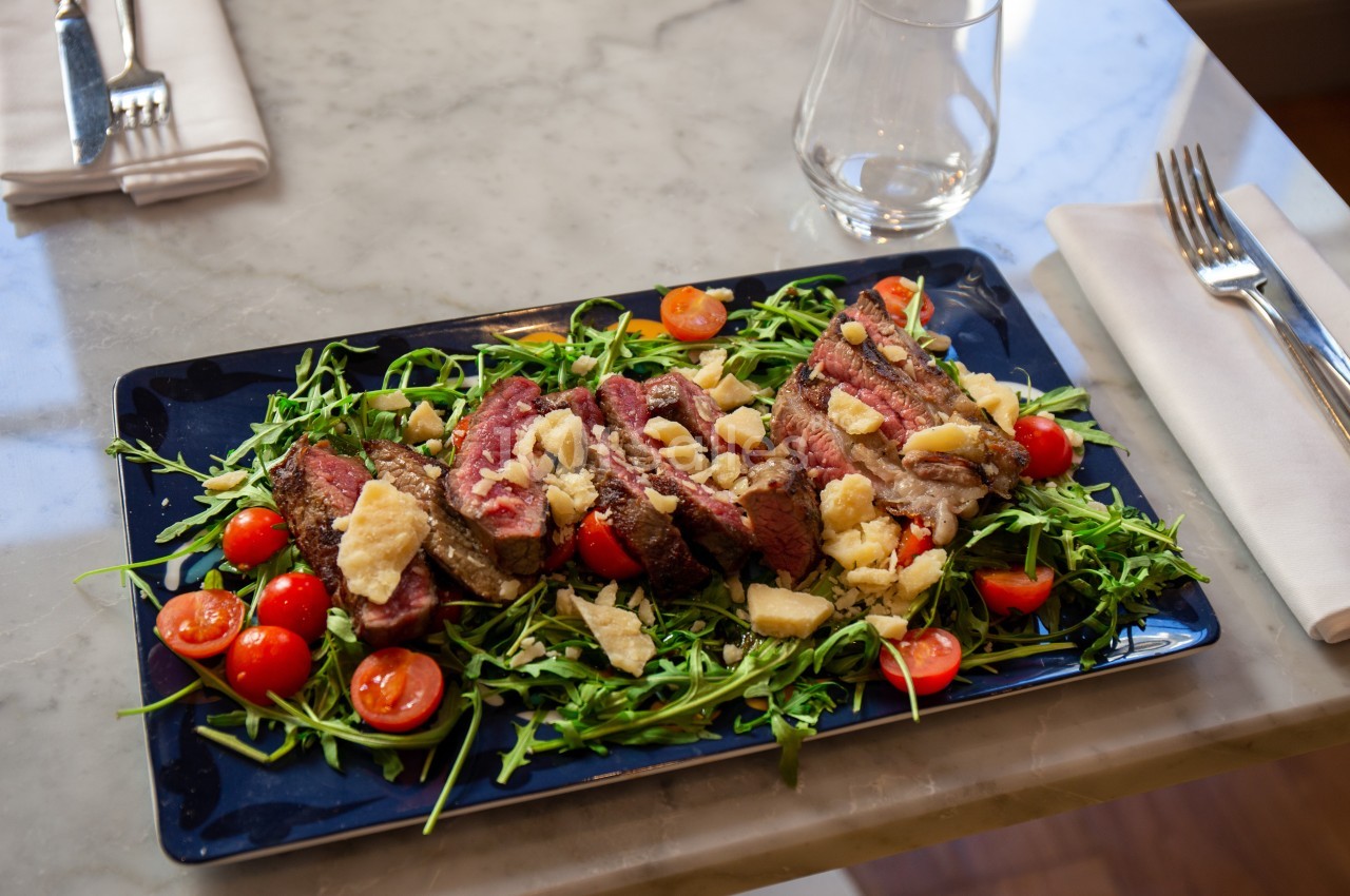 Assiette de viande grillée garnie de roquette, tomates cerises et copeaux de fromage, servie sur une table en marbre.