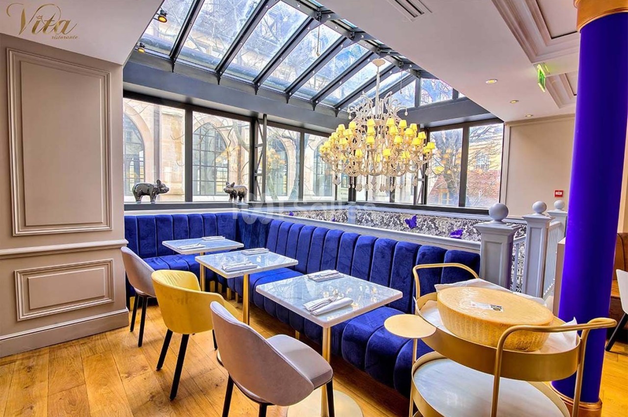 Salle de restaurant lumineuse avec verrière, banquettes bleues, chaises modernes et lustre élégant.