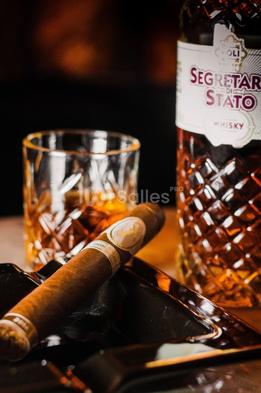 Un cigare posé sur un cendrier noir, avec un verre de whisky et une bouteille en arrière-plan.