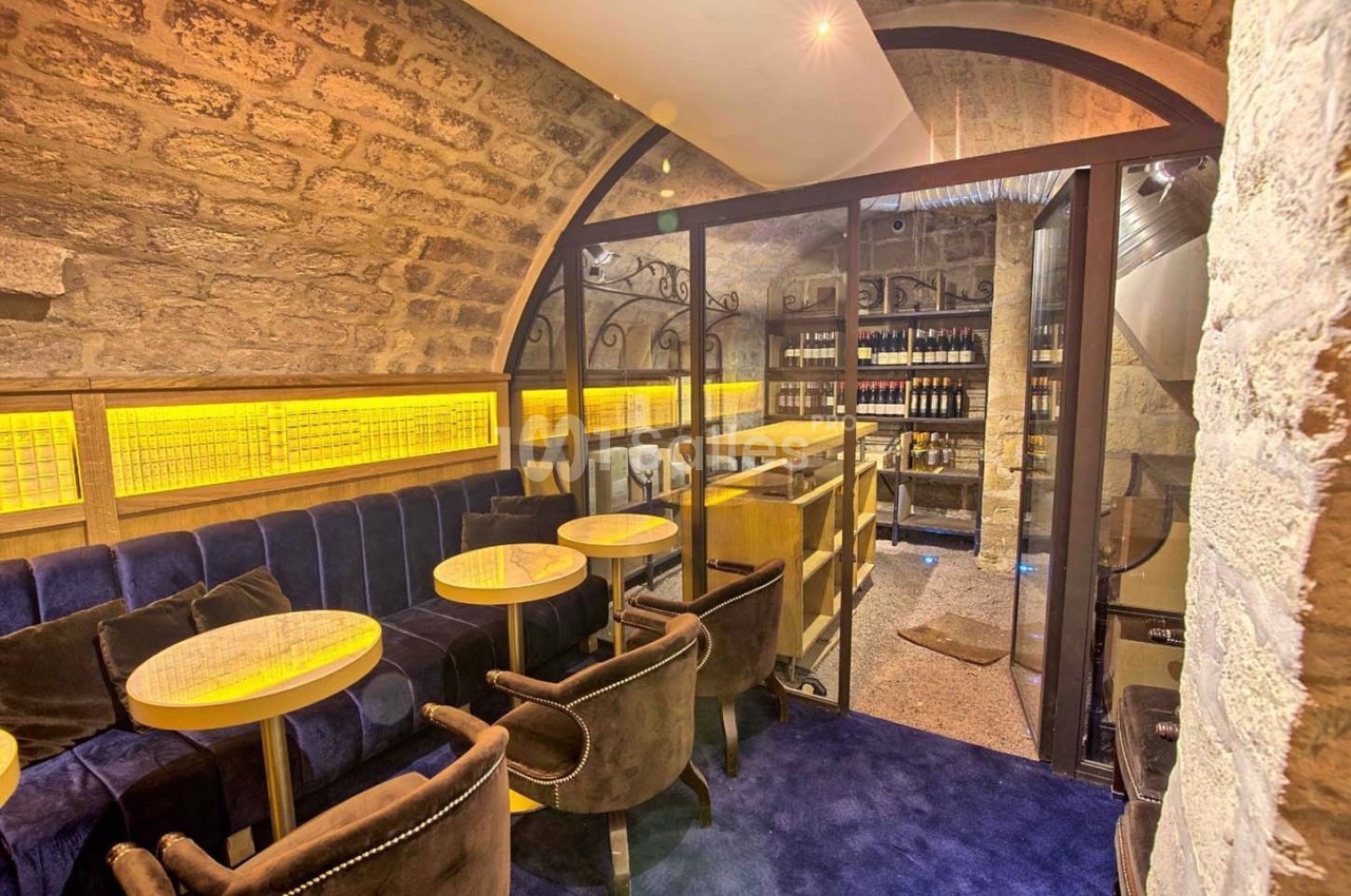 Salle voûtée en pierre avec tables, chaises en velours et étagères de bouteilles de vin derrière une cloison vitrée.