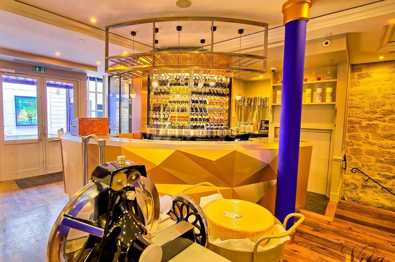 Intérieur d'une boutique avec un comptoir doré, une cave à vin vitrée, une trancheuse et une meule de fromage.