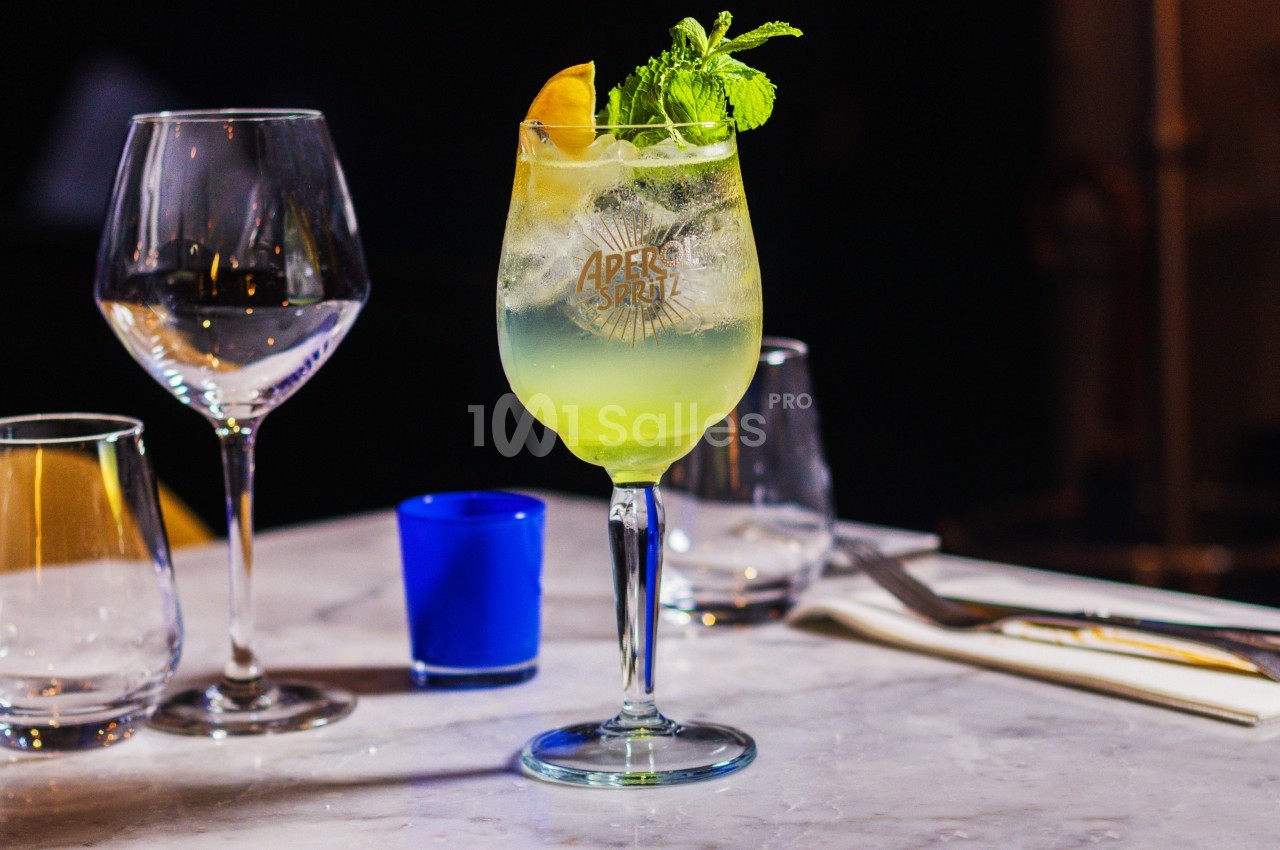 Verre de cocktail jaune décoré de menthe et citron sur une table en marbre, entouré de verres et couverts.