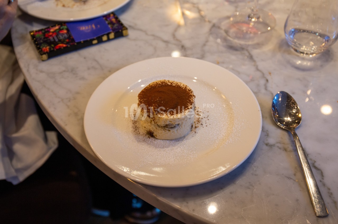 Dessert tiramisu saupoudré de cacao servi sur une assiette blanche posée sur une table en marbre.