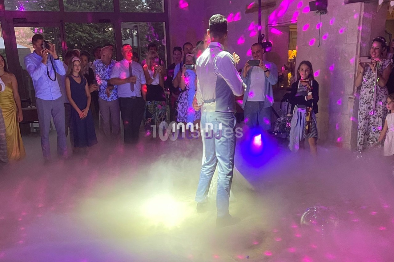 Un homme en costume danse au centre d'une piste entourée d'invités, éclairée par des lumières colorées et de la fumée.