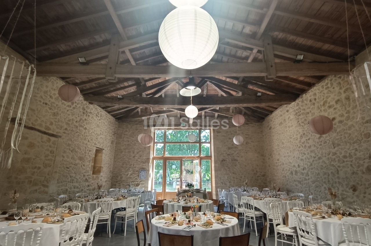 Salle de réception en pierre avec tables décorées pour un événement, éclairée par des lanternes suspendues.