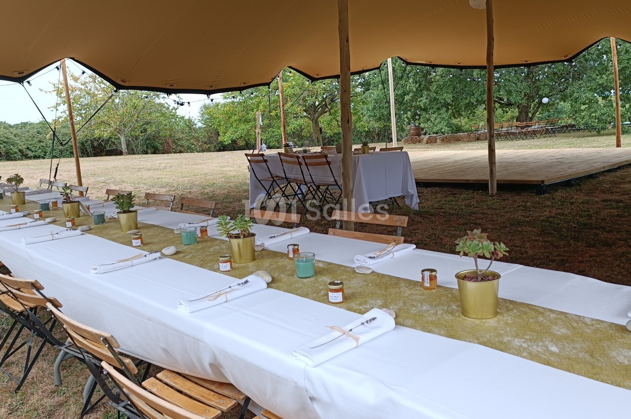 Tables dressées avec nappes blanches, plantes en pot et bougies, sous une tente dans un espace extérieur verdoyant.