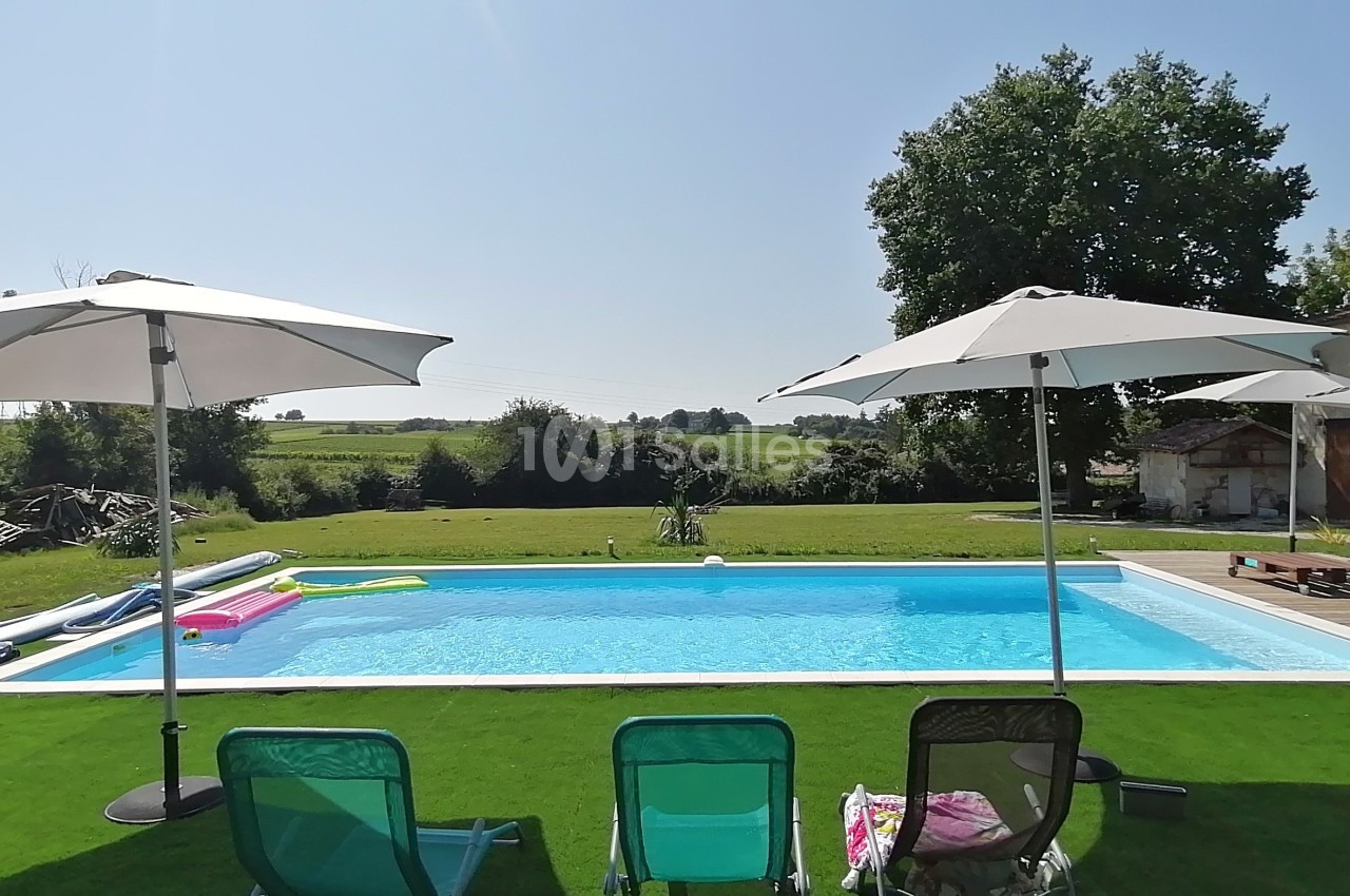 Piscine extérieure entourée de chaises longues, parasols et pelouse, avec vue sur la campagne en arrière-plan.