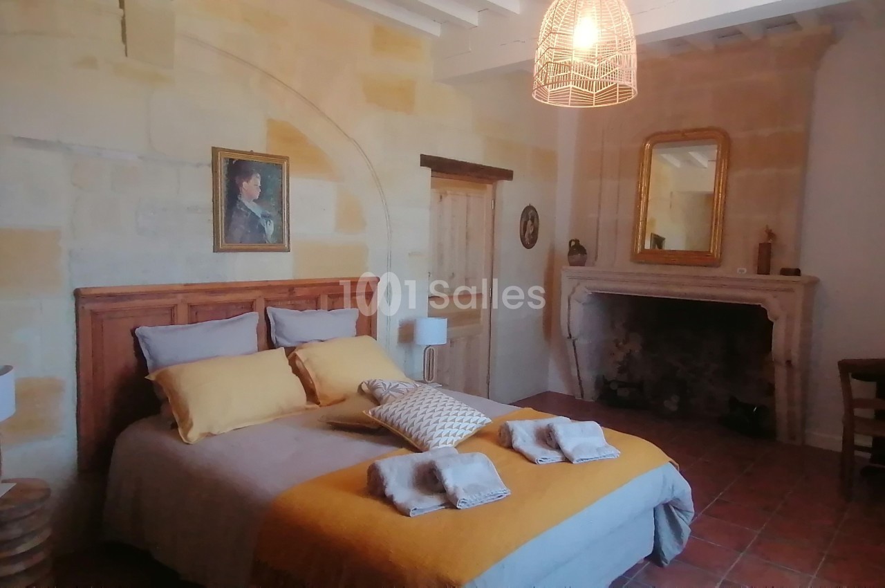 Chambre chaleureuse avec lit double, murs en pierre, cheminée ancienne, miroir doré et éclairage suspendu.
