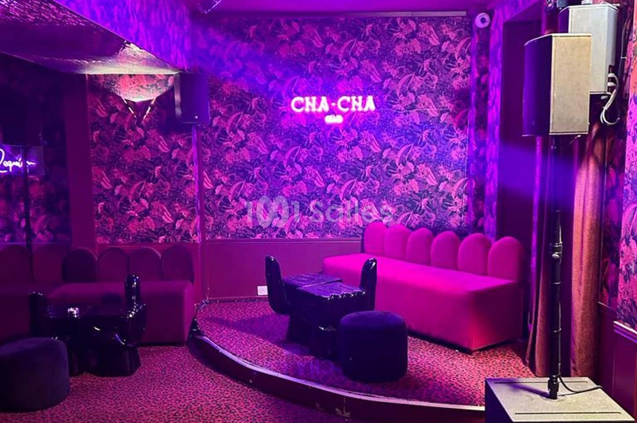 Salle colorée avec canapé rose, murs à motifs, éclairage violet et enseigne lumineuse ’CHA-CHA’.