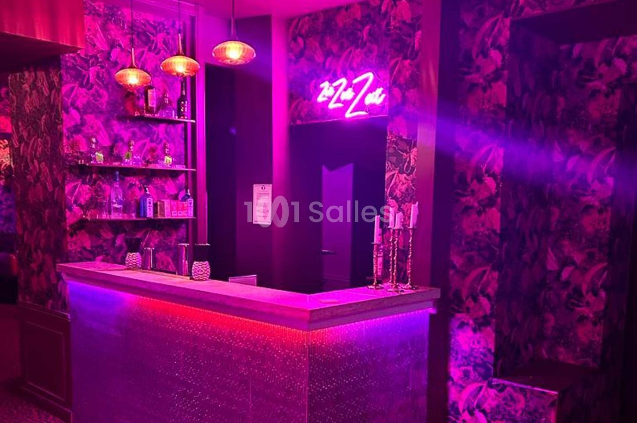 Comptoir de bar éclairé par des lumières roses et violettes, avec décor floral et enseigne néon au mur.