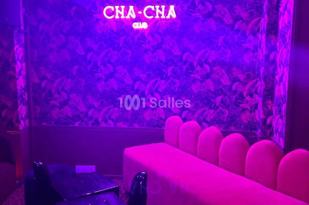 Banquette rose et fauteuil noir dans une pièce éclairée par une lumière violette avec un néon ’CHA-CHA Club’ au mur.