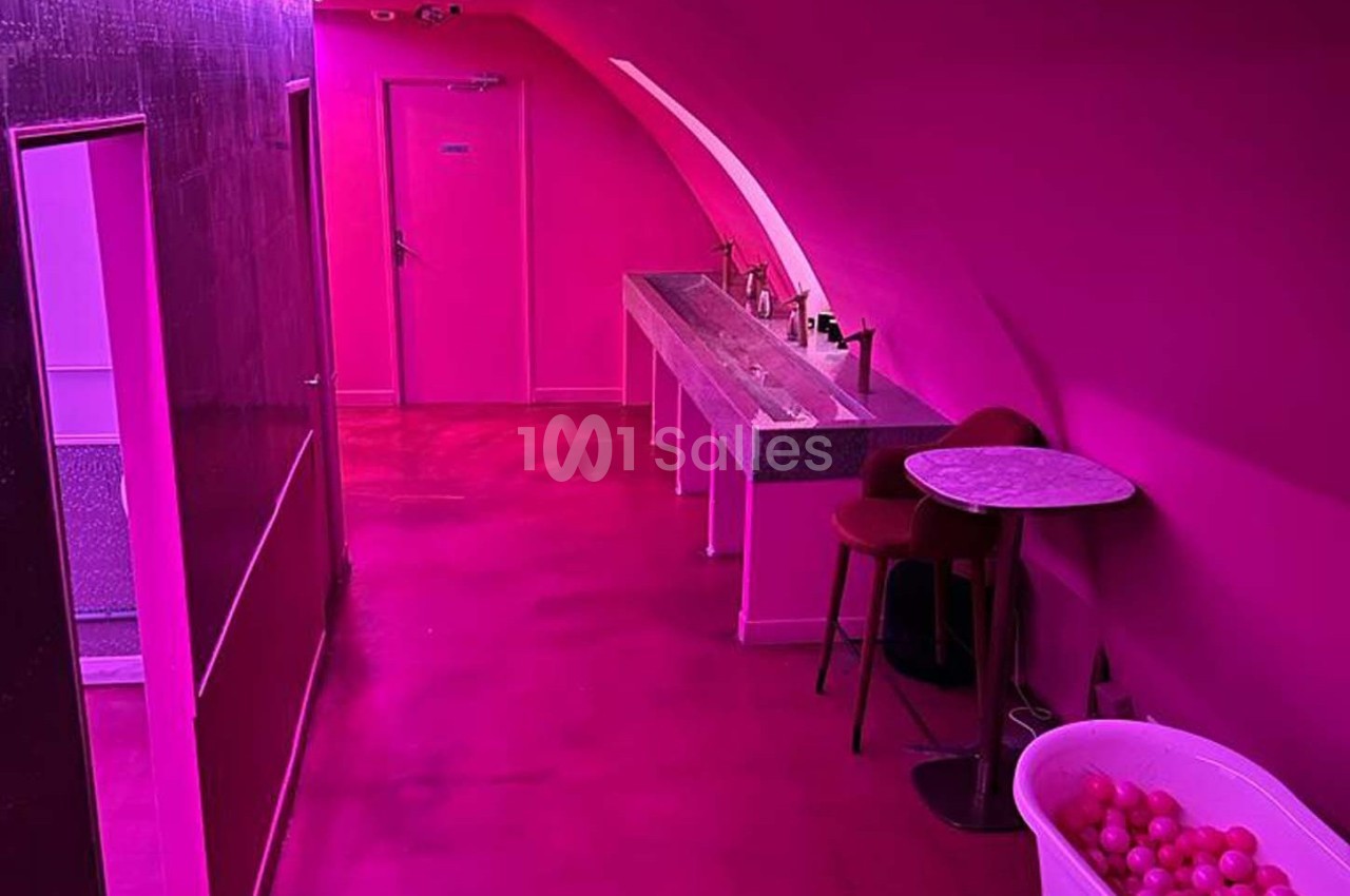 Couloir intérieur aux murs et sol roses, avec un lavabo, une table, une chaise et une bassine remplie de balles.