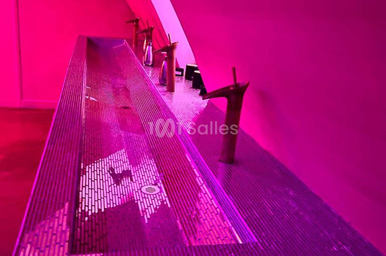 Lavabo collectif moderne avec robinetterie design, éclairé par une lumière rose vive.