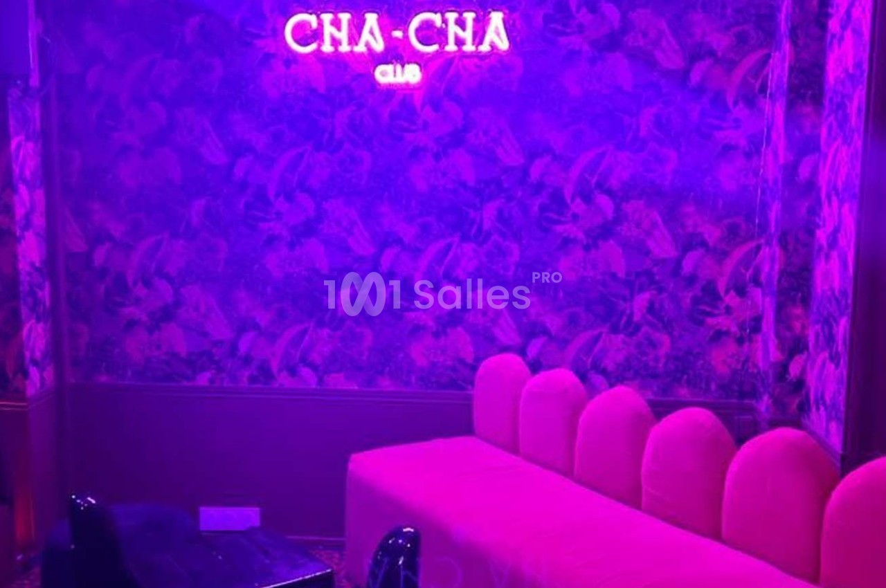 Banquette rose et mur à motifs végétaux éclairés par une lumière violette, avec un néon ’CHA-CHA CLUB’.