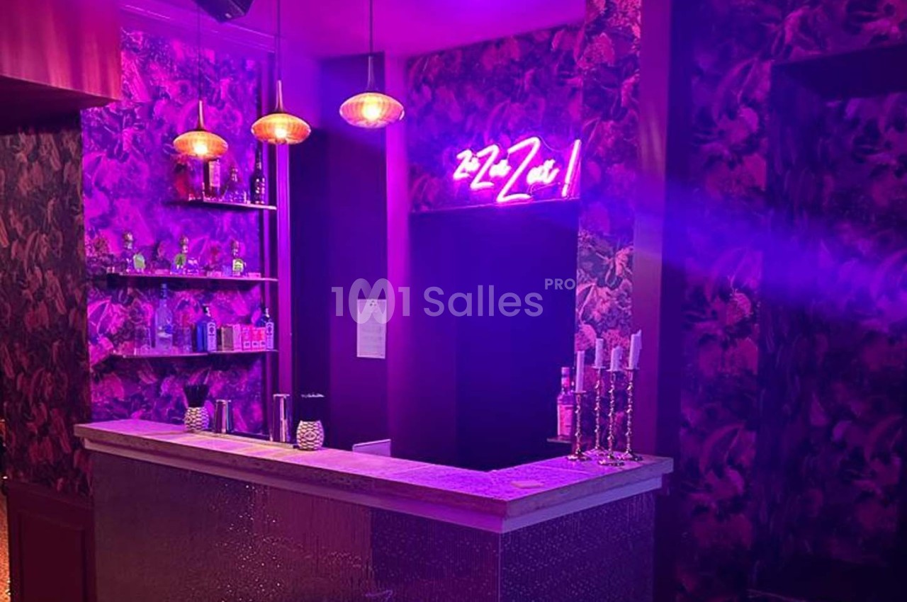 Bar avec comptoir éclairé, étagères de bouteilles, murs décorés et enseigne néon rose dans une ambiance tamisée.