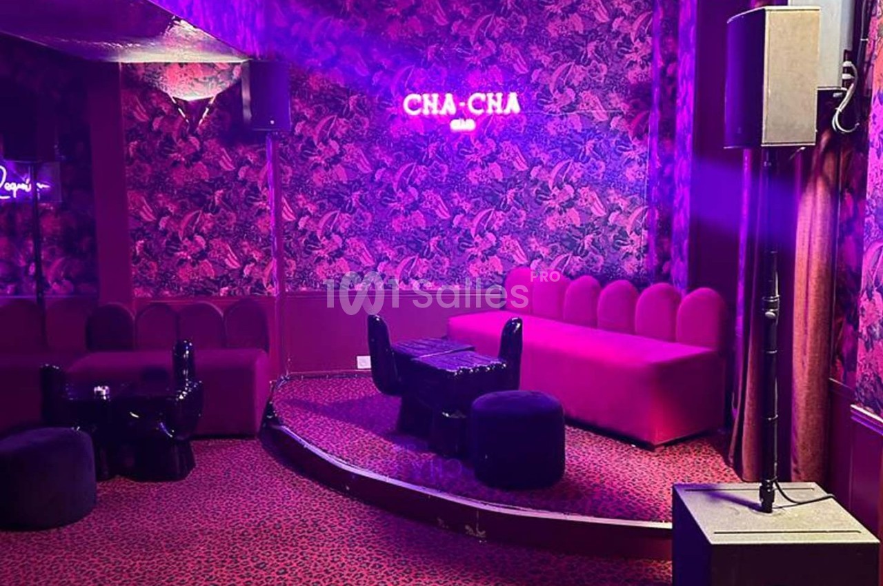Salle avec banquettes rouges, table basse noire, éclairage tamisé et mur à motif floral, enseigne ’CHA-CHA’ lumineuse.