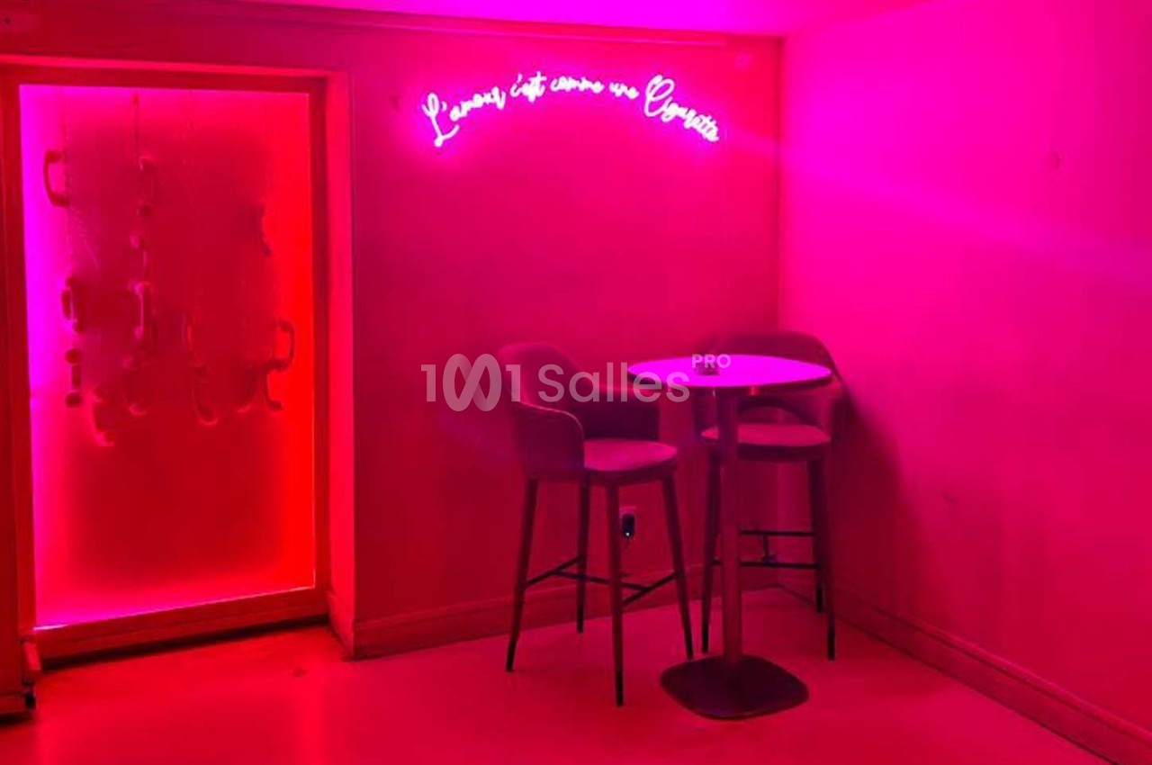 Deux chaises hautes et une table ronde dans une pièce éclairée par une lumière rose avec un néon au mur.