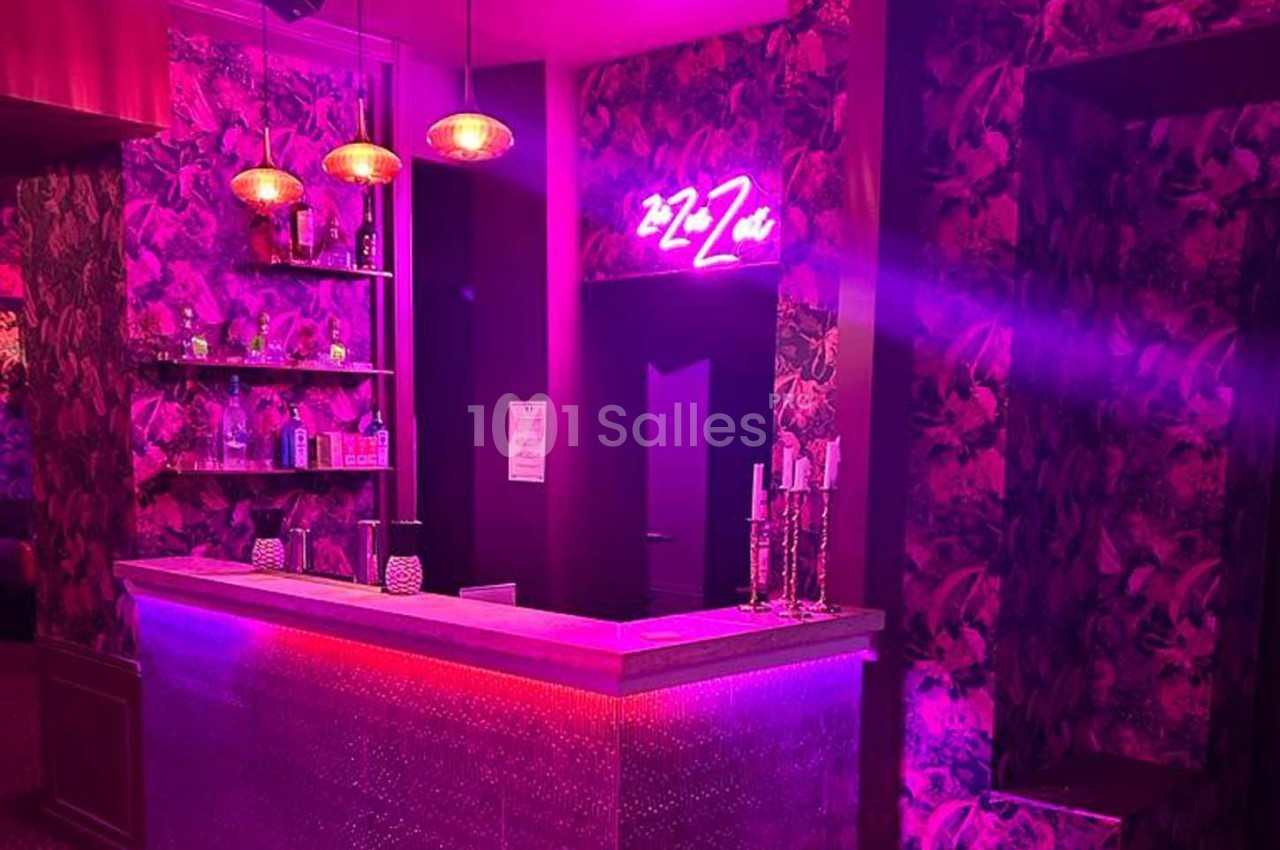 Bar avec éclairage rose et violet, décor floral, étagères avec bouteilles, et enseigne lumineuse au mur.