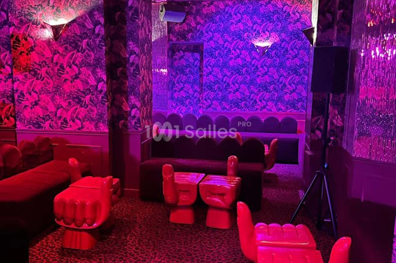 Salle au décor rétro avec fauteuils en forme de mains rouges, murs à motifs et éclairage rose-violet tamisé.