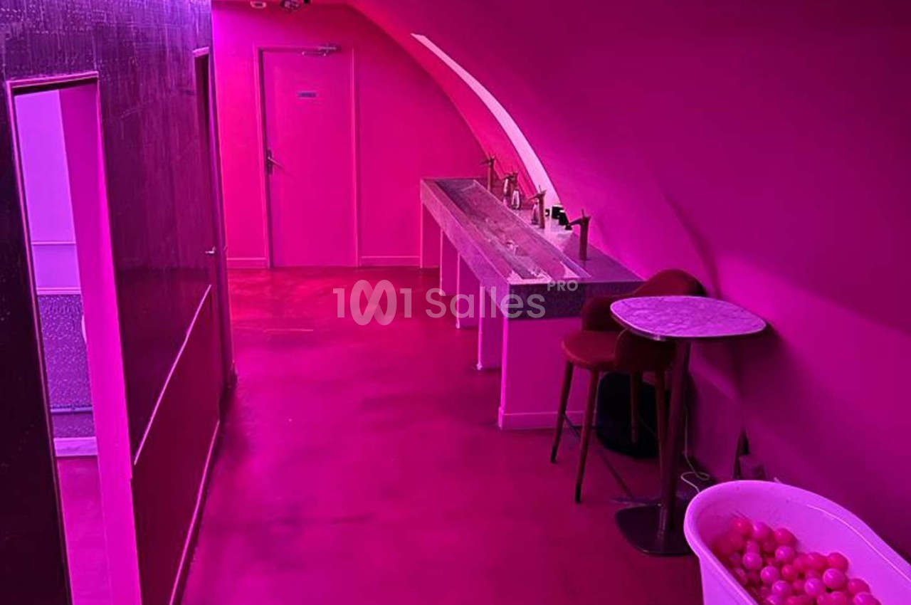 Salle aux murs et sol roses avec un lavabo, une table, une chaise et une baignoire remplie de balles colorées.