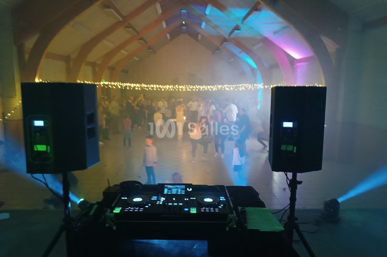 Vue depuis une table de mixage avec des enceintes, éclairage coloré et une foule dansante dans une salle festive.