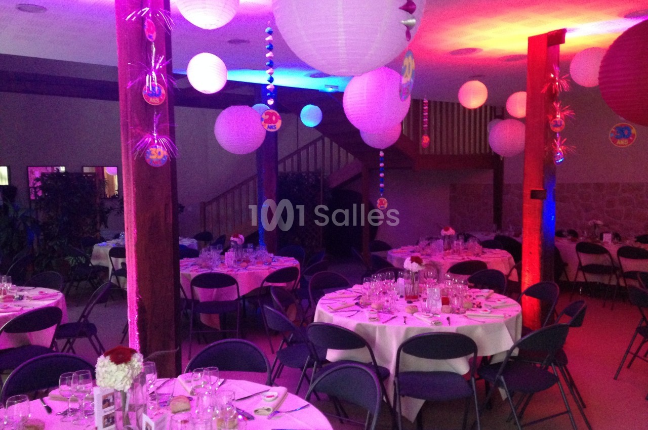 Salle décorée pour un événement, avec tables rondes dressées, lanternes suspendues et éclairage coloré.