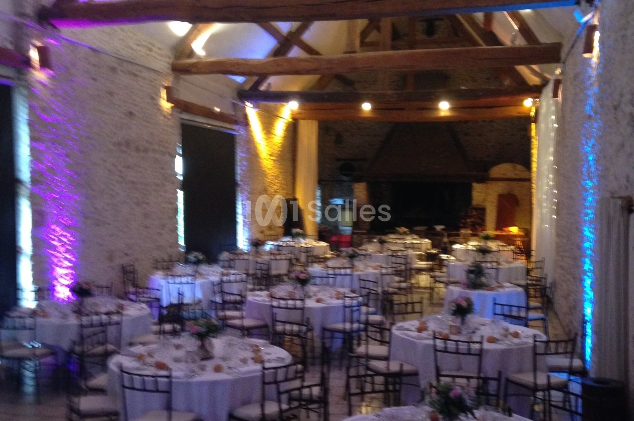 Salle de réception avec des tables rondes dressées, décorée de poutres apparentes et éclairages colorés.