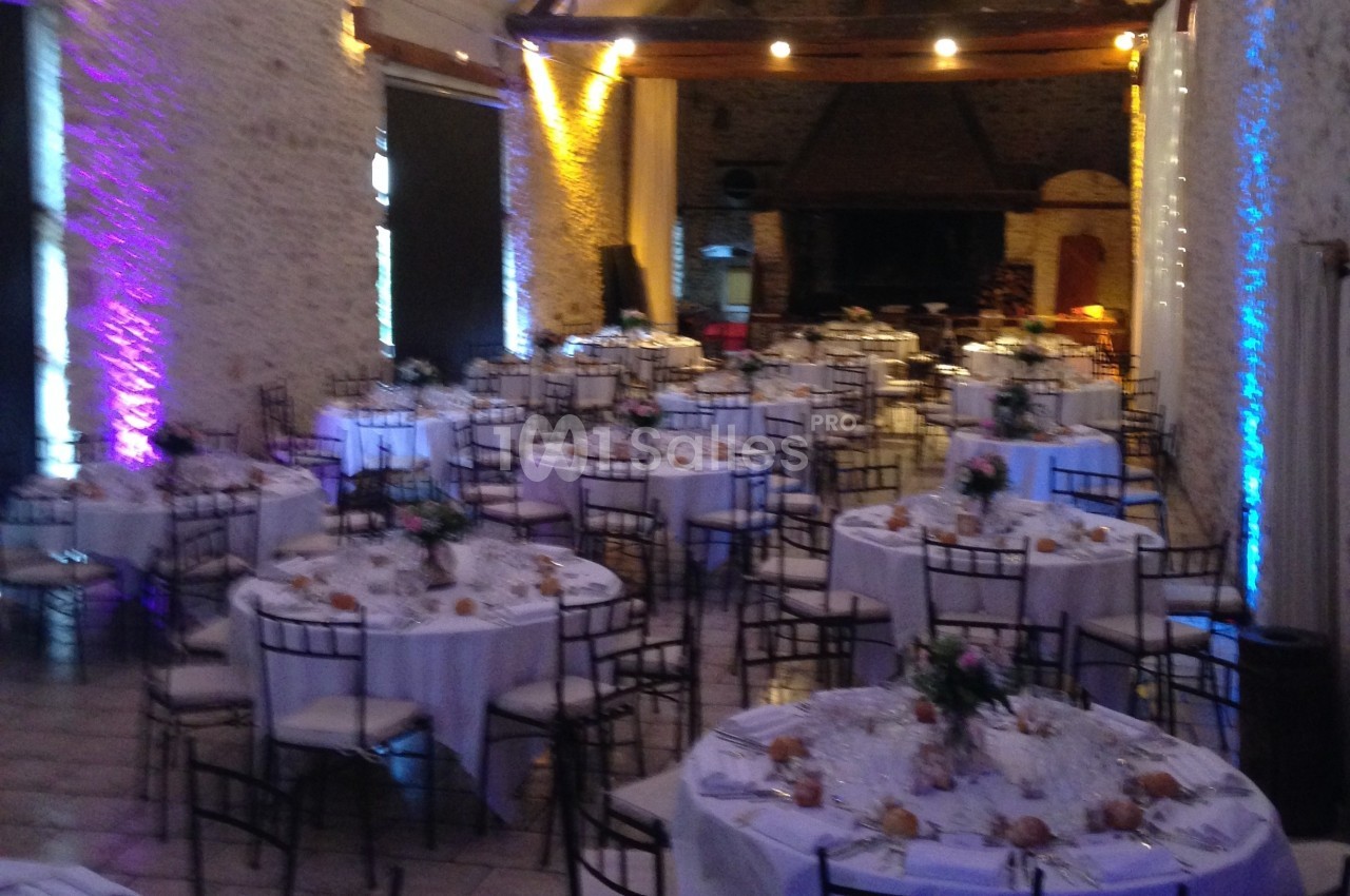 Salle de réception avec des tables rondes dressées, nappes blanches, chaises en métal et éclairage coloré.