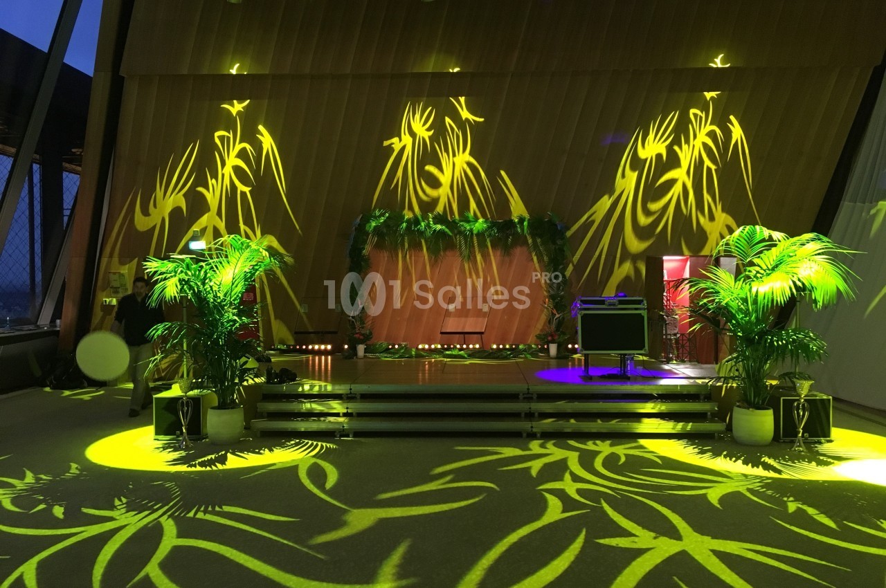 Scène décorée de plantes et éclairée par des motifs lumineux jaunes dans une salle moderne avec de grandes fenêtres.