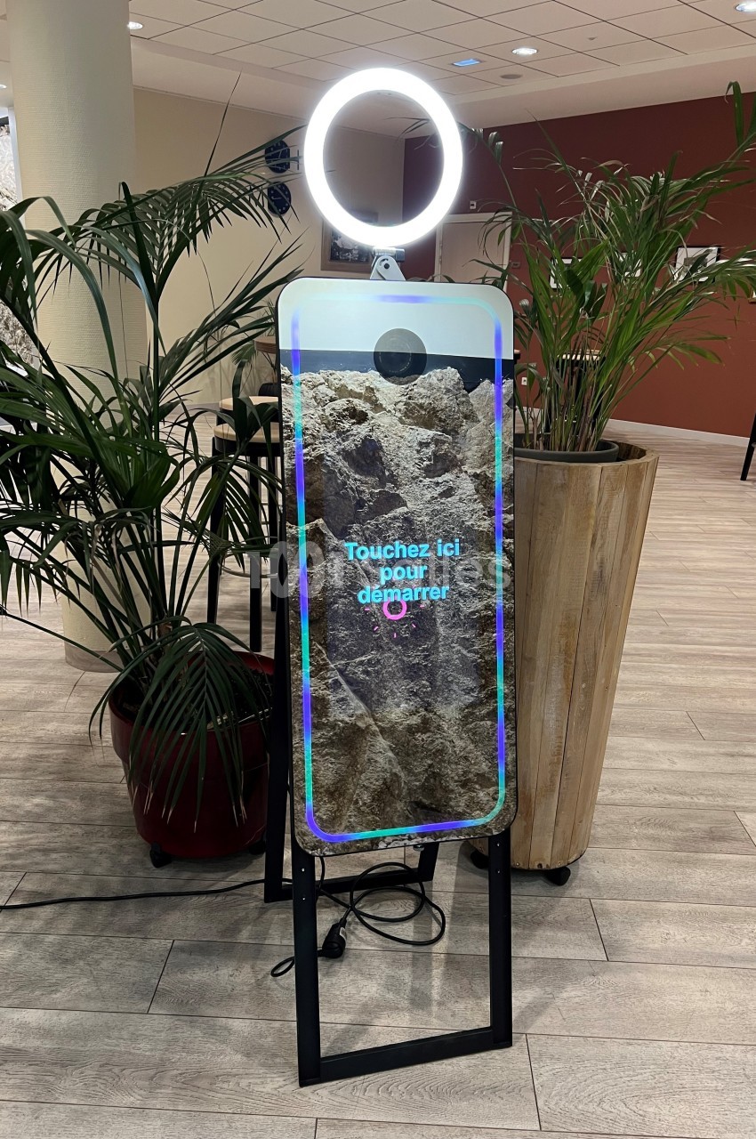 Borne photo interactive avec écran tactile et anneau lumineux, installée dans un espace intérieur avec des plantes…