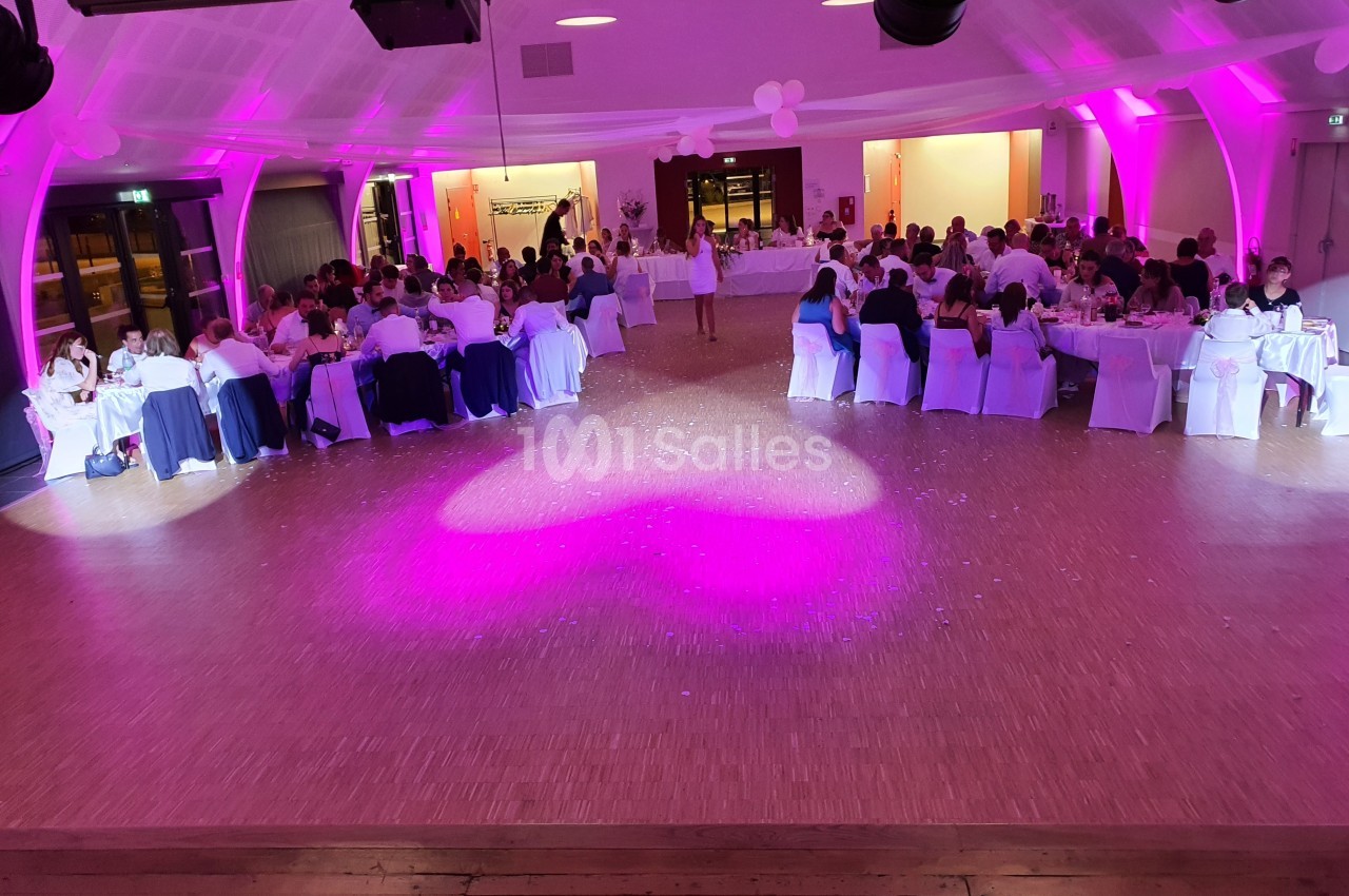 Salle de réception éclairée en rose, avec des invités assis à des tables rondes lors d'un événement festif.