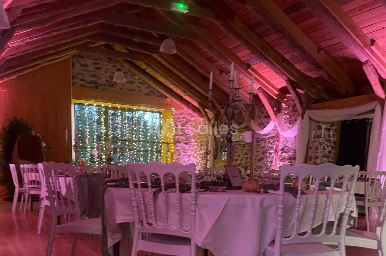 Salle de réception décorée avec des guirlandes lumineuses, des tables dressées et un plafond en poutres apparentes.