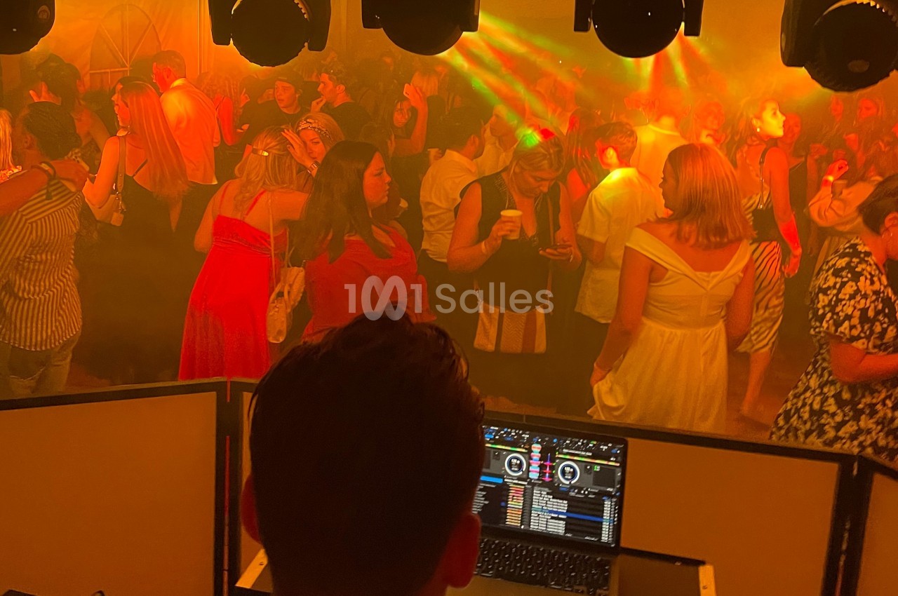 Vue depuis la cabine d’un DJ sur une piste de danse bondée, éclairée par des lumières colorées et une ambiance festive.