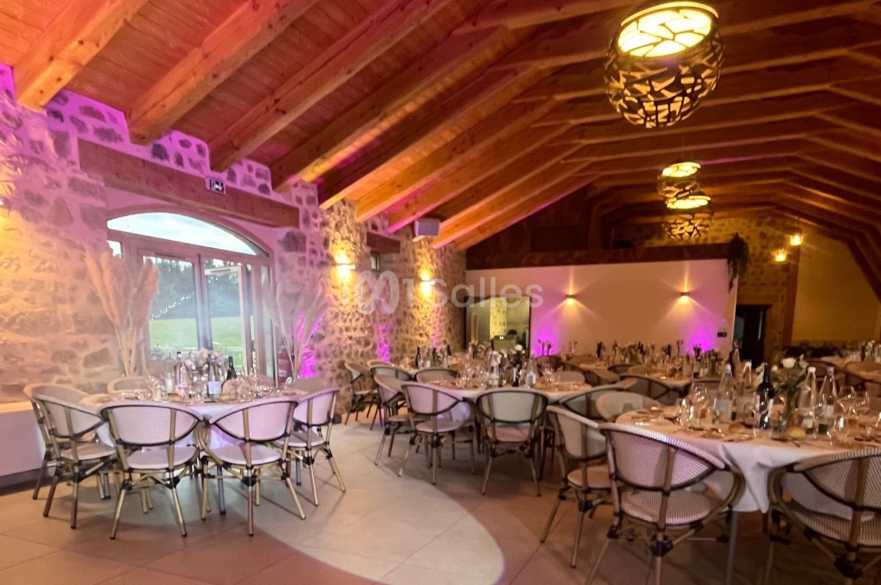 Salle de réception en pierre avec plafond en bois, tables rondes dressées et éclairage chaleureux.