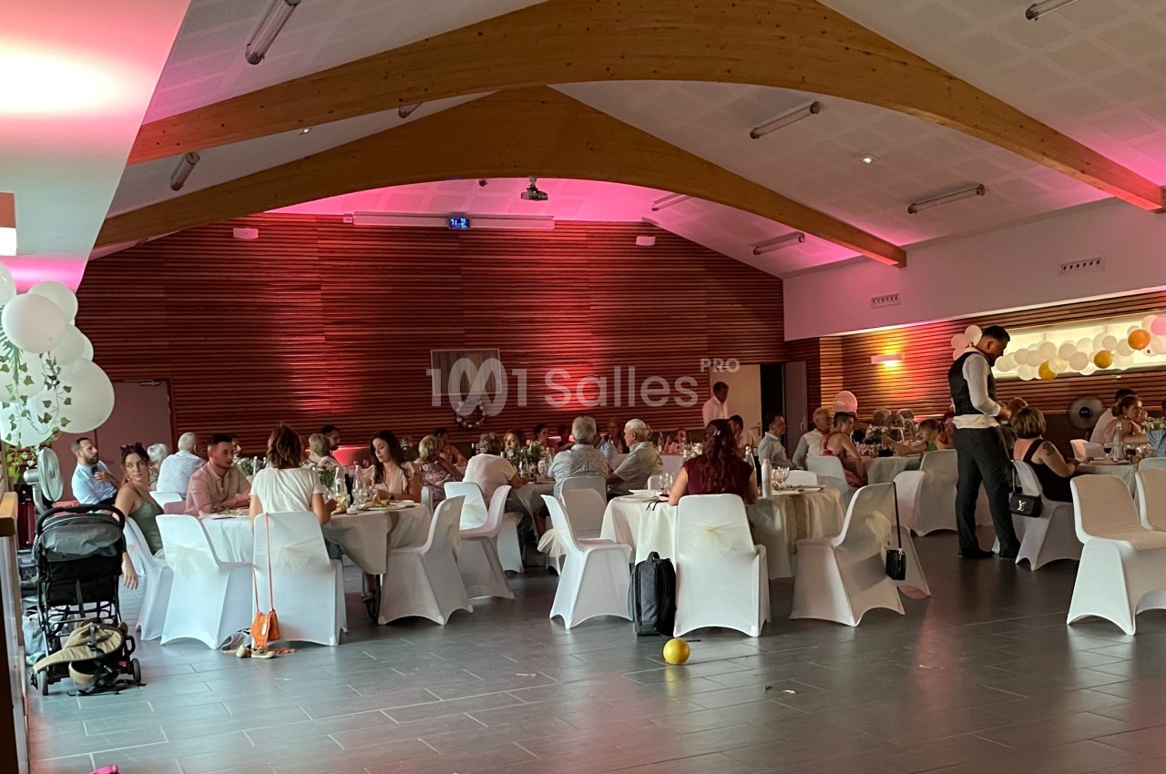 Salle décorée avec des ballons et éclairages roses, accueillant des invités assis à des tables pour un événement festif.