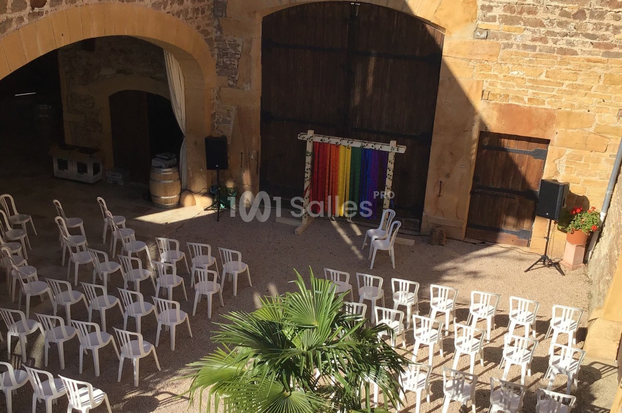 Cour intérieure ensoleillée avec des rangées de chaises blanches disposées face à une scène décorée de rideaux colorés.