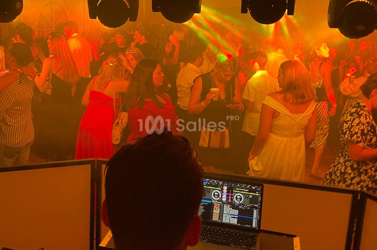 Vue depuis la cabine d’un DJ sur une piste de danse bondée, éclairée par des lumières colorées et une ambiance festive.