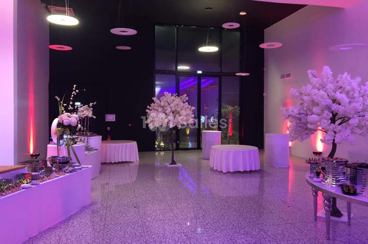 Salle de réception décorée avec des tables rondes drapées de blanc, des arbres artificiels et un éclairage coloré.
