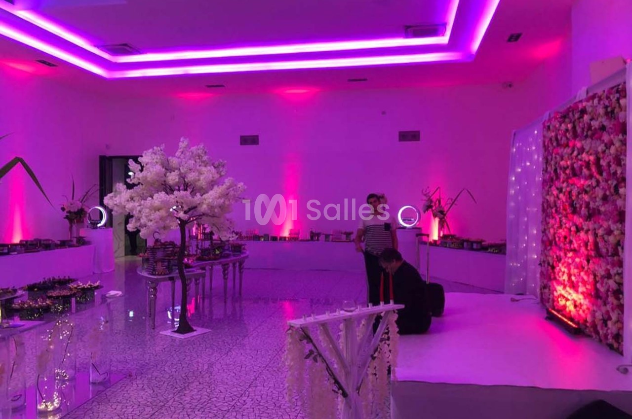 Salle décorée avec des lumières violettes, un arbre artificiel blanc et un mur floral, avec deux personnes présentes.