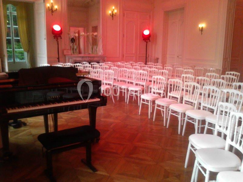 Salle élégante avec parquet, rangées de chaises blanches, un piano à queue noir et des projecteurs rouges allumés.