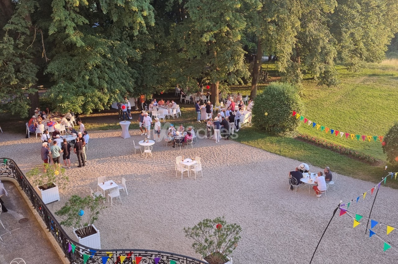 Vue d'une réception en plein air avec des invités rassemblés dans une cour, entourée de verdure et décorée de guirlandes…