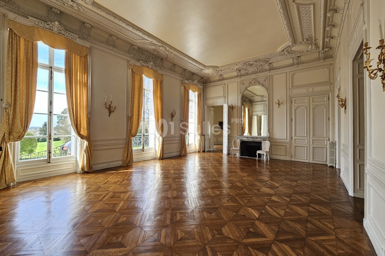 Grande salle lumineuse avec parquet en bois, hauts plafonds ornés, larges fenêtres et rideaux dorés donnant sur un jardin.