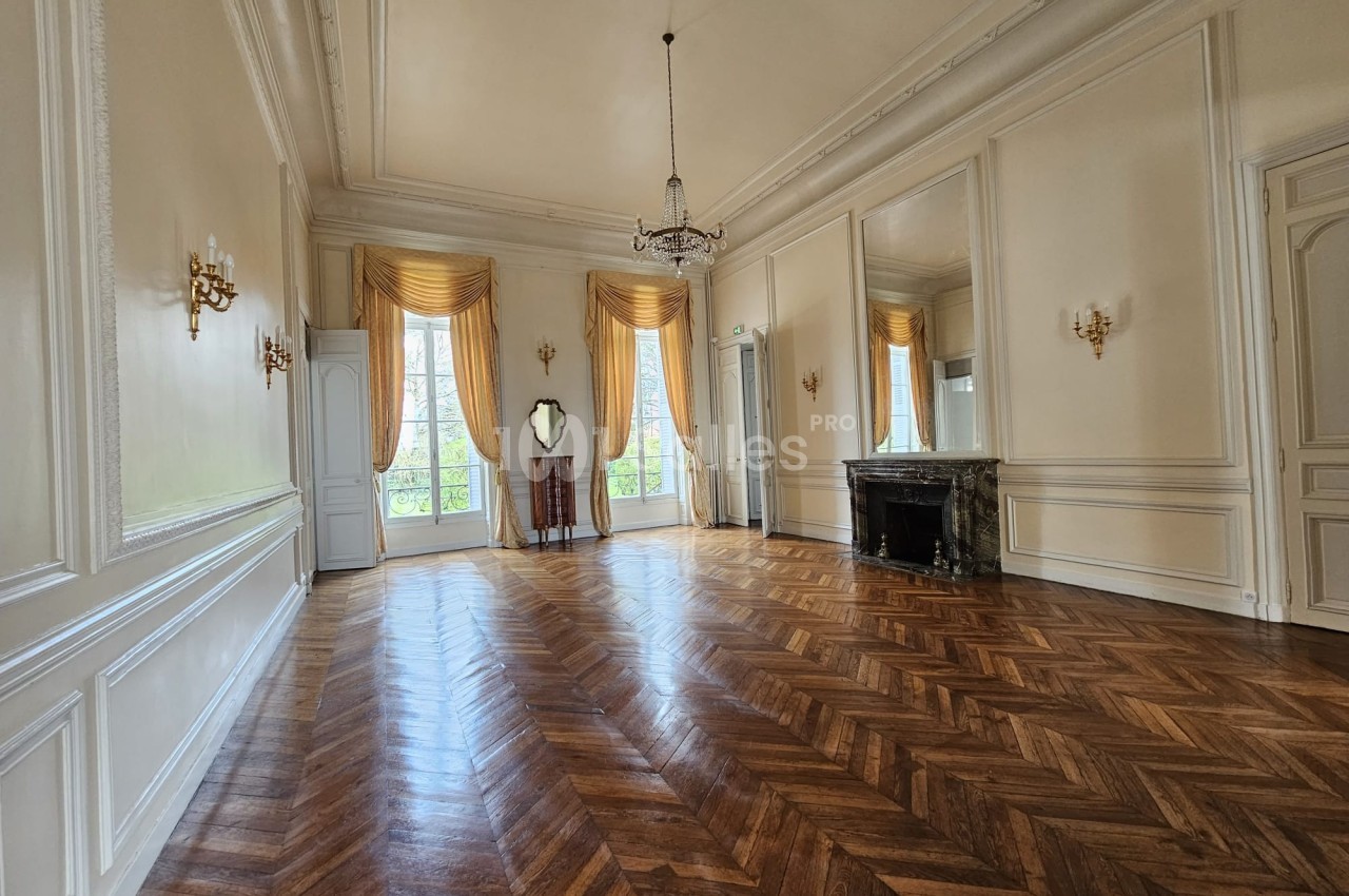 Salle lumineuse avec parquet en chevrons, grandes fenêtres, rideaux dorés, cheminée en marbre et miroirs muraux.