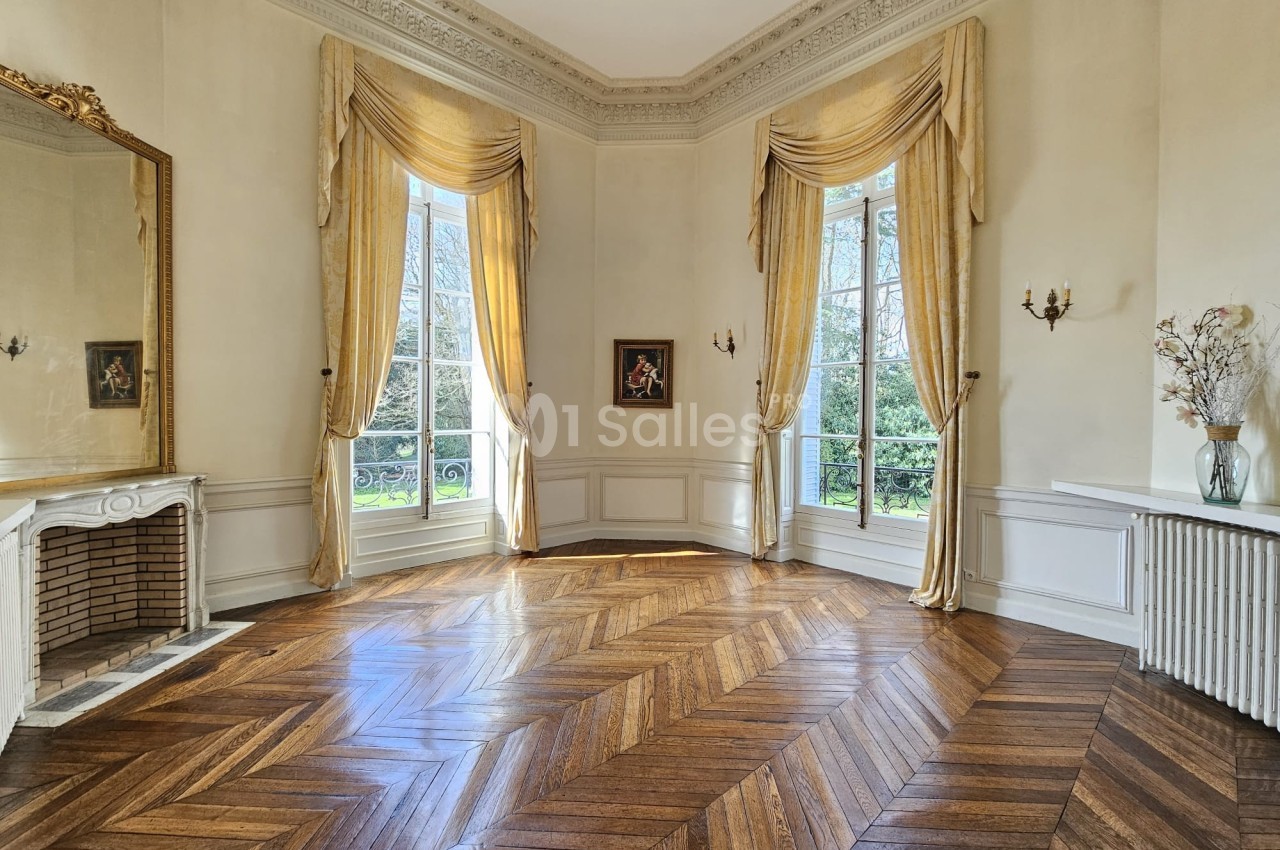 Salon lumineux avec parquet en chevrons, grandes fenêtres encadrées de rideaux dorés, cheminée et miroir décoratif.